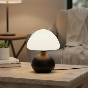 May include: A contemporary table lamp featuring a white, mushroom-shaped lampshade atop a dark brown base. The lamp sits on a wooden table. The design is minimalist. The background is neutral-toned.