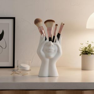 Puede incluir: Un soporte blanco de cerámica para brochas de maquillaje con forma de rostro con manos. El soporte contiene varios pinceles de maquillaje con cerdas marrones y mangos de oro rosa. El soporte se encuentra sobre una superficie blanca con una pequeña planta y un cuadro en el fondo.