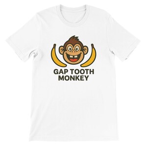 Puede incluir: Camiseta blanca con un diseño de mono de dibujos animados con una sonrisa con huecos entre los dientes, flanqueado por dos plátanos amarillos. El texto "GAP TOOTH MONKEY" está impreso en marrón debajo. La camiseta es de material suave.