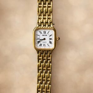 Puede incluir: Un reloj Croton de cuarzo dorado con una esfera rectangular y una correa de eslabones dorados a juego. La esfera del reloj es blanca con números romanos y agujas negras. La correa mide unos 18 cm de largo.