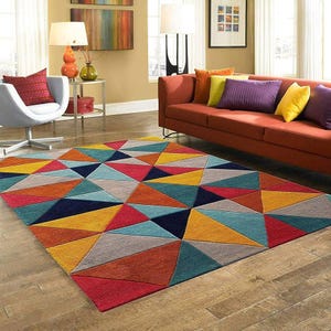 May include: A colorful geometric area rug with a modern design. The rug features a pattern of triangles in shades of red, orange, yellow, blue, and gray. The rug is placed on a hardwood floor in a living room setting.