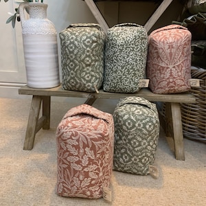 May include: A selection of fabric doorstops in shades of green and red, featuring floral patterns. The doorstops are cylindrical with a folded top and a small tag attached.