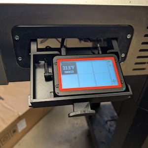 Könnte beinhalten: Ein digitaler Smoker-Controller mit schwarzem Rahmen und rotem Rand. Der Bildschirm zeigt die Temperatur als 33,5°F an. Das Wort "SMOKER" ist ebenfalls auf dem Bildschirm zu sehen. Der Controller ist an einem Metallgerät montiert.