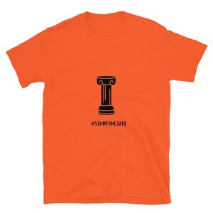 Pode incluir: Camiseta laranja brilhante com um gráfico preto de uma coluna clássica e o texto "#STANDINGTALL". A camiseta é feita de um material macio. O design está centrado na frente da camiseta.