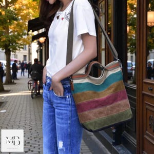 May include: A striped canvas tote bag with a shoulder strap. The bag has horizontal stripes in green, brown, burgundy, and beige. The bag is carried by a person wearing a white t-shirt and blue jeans.