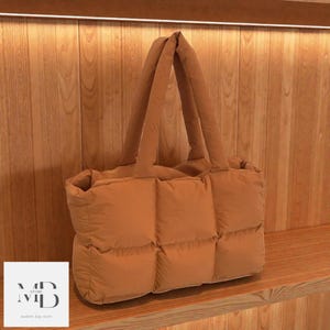 Peut inclure: Un sac fourre-tout matelassé de couleur beige avec deux poignées rembourrées. Le sac a une forme rectangulaire avec une série de sections rembourrées de forme carrée. Le sac est présenté sur une étagère en bois.