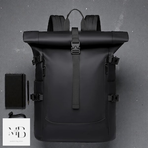 May include: Black roll-top backpack with adjustable straps and a buckle closure. The bag has multiple side pockets and a handle at the top. A black notebook, pen, and a small bag with text are next to the backpack.