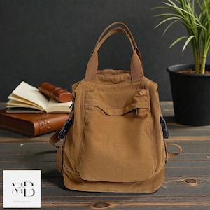 Può includere: Una borsa tote in tela marrone con manico superiore e tasca frontale. La borsa ha una forma rettangolare ed è realizzata in materiale resistente. La borsa è appoggiata su una superficie di legno. La borsa ha un logo nell'angolo in basso a sinistra.