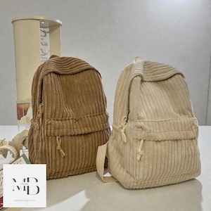 Puede incluir: Dos mochilas de pana en tonos neutros. Una es de color marrón cálido, la otra de color crema suave. Ambas tienen un bolsillo frontal, una solapa superior y correas ajustables. Las mochilas se exhiben sobre una superficie blanca.