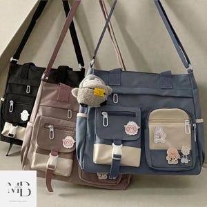 Puede incluir: Tres bolsos de hombro en negro, malva y gris azulado, cada uno con múltiples bolsillos delanteros y un cierre de hebilla blanco. Los bolsos tienen parches decorativos y un pequeño llavero de pingüino de peluche. Los bolsos tienen un diseño casual.