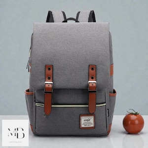 Può includere: Zaino grigio con dettagli in pelle marrone, tra cui cinghie e tasche laterali. Presenta una chiusura a patta con fibbie e una piccola etichetta. Un pomodoro è a destra dello zaino.