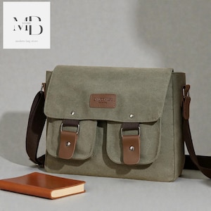 May include: Olive green canvas messenger bag with two front pockets and brown leather accents. The bag has a brown shoulder strap and a flap closure. A brown leather-bound notebook rests in front of the bag.