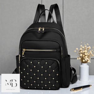 Puede incluir: Mochila negra con cremalleras doradas y un bolsillo frontal con tachuelas. La mochila tiene dos bolsillos laterales y correas ajustables. El texto "MD STORE modern bag store" es visible en la esquina inferior izquierda.