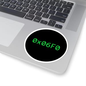 May include: A round sticker with a black background and the text "0x06F0" in green. The sticker is placed on a silver laptop, with the keyboard visible in the background.