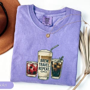 Puede incluir: Una camiseta violeta Comfort Colors con un gr&aacute;fico de tres vasos de bebidas. El vaso central tiene el texto "Brew. Travel. Repeat." Un collar dorado y un sombrero marr&oacute;n est&aacute;n junto a la camiseta, junto con una planta seca.