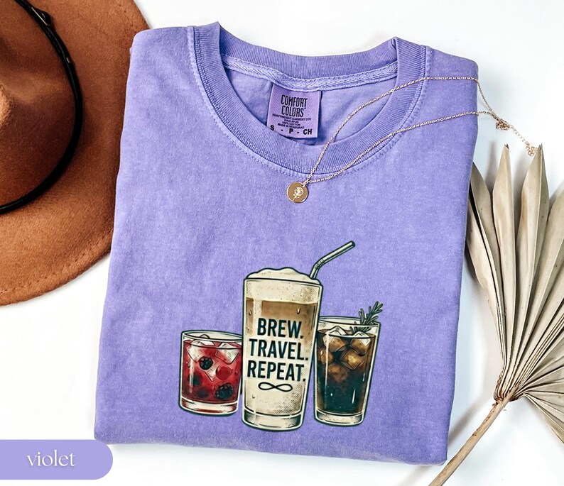 Puede incluir: Una camiseta violeta Comfort Colors con un gr&aacute;fico de tres vasos de bebidas. El vaso central tiene el texto "Brew. Travel. Repeat." Un collar dorado y un sombrero marr&oacute;n est&aacute;n junto a la camiseta, junto con una planta seca.