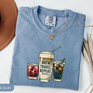 Puede incluir: Una camiseta azul vaquero Comfort Colors con un gr&aacute;fico de tres bebidas: un c&oacute;ctel, una bebida de caf&eacute; con el texto "BREW. TRAVEL. REPEAT." y una bebida oscura. Un collar dorado est&aacute; sobre la camiseta.