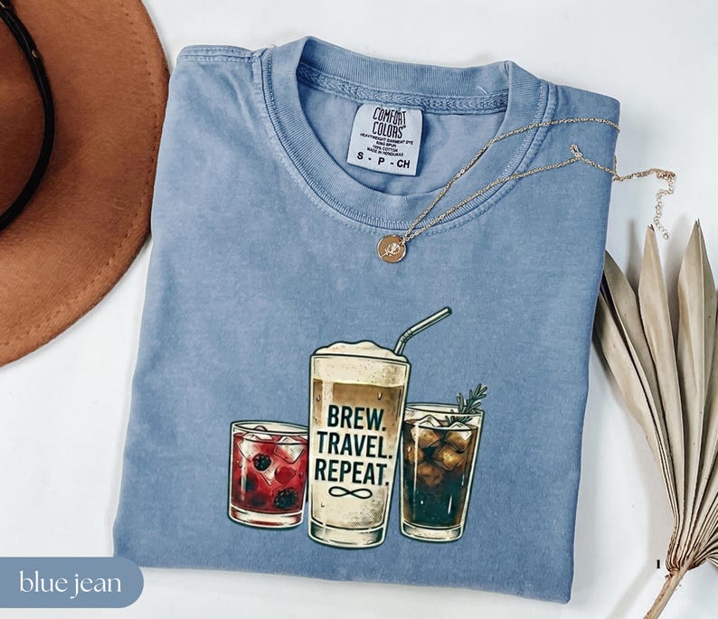 Puede incluir: Una camiseta azul vaquero Comfort Colors con un gr&aacute;fico de tres bebidas: un c&oacute;ctel, una bebida de caf&eacute; con el texto "BREW. TRAVEL. REPEAT." y una bebida oscura. Un collar dorado est&aacute; sobre la camiseta.