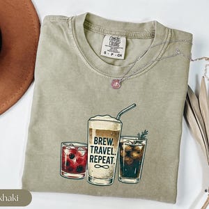 Puede incluir: Camiseta color caqui con un gr&aacute;fico de tres bebidas: un c&oacute;ctel, una bebida de caf&eacute; con el texto "Brew. Travel. Repeat.", y una cola. La camiseta tiene cuello redondo y mangas cortas. Un collar plateado con un colgante rosa est&aacute; sobre la camiseta.