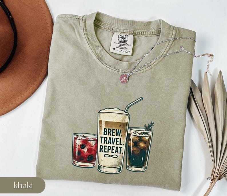 Puede incluir: Camiseta color caqui con un gr&aacute;fico de tres bebidas: un c&oacute;ctel, una bebida de caf&eacute; con el texto "Brew. Travel. Repeat.", y una cola. La camiseta tiene cuello redondo y mangas cortas. Un collar plateado con un colgante rosa est&aacute; sobre la camiseta.
