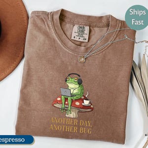 May include: Espresso-colored t-shirt with a graphic of a green frog wearing headphones and using a laptop, sitting on a mushroom. The text "ANOTHER DAY, ANOTHER BUG" is below the graphic. The shirt has a crew neck and short sleeves.