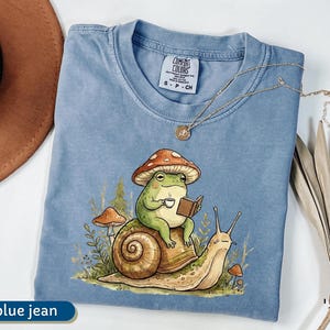 May include: A blue jean Comfort Colors t-shirt featuring a whimsical illustration of a green frog wearing a mushroom hat, reading a book, and holding a cup of coffee while riding a snail. A gold necklace is draped across the shirt.