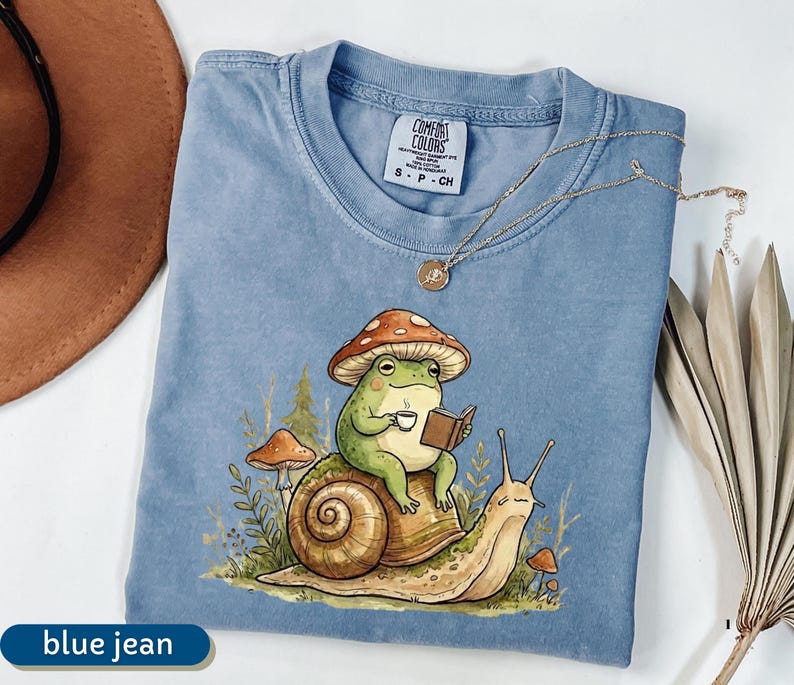 May include: A blue jean Comfort Colors t-shirt featuring a whimsical illustration of a green frog wearing a mushroom hat, reading a book, and holding a cup of coffee while riding a snail. A gold necklace is draped across the shirt.
