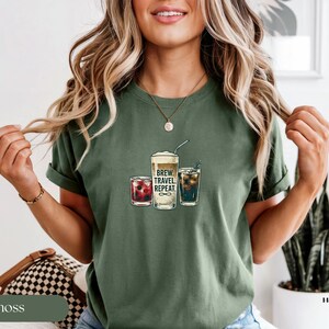 Puede incluir: Camiseta verde musgo con un gr&aacute;fico de tres bebidas: un caf&eacute; espumoso, un c&oacute;ctel rojo y una bebida oscura con hielo. El texto "BREW. TRAVEL. REPEAT." est&aacute; impreso en el caf&eacute;. La modelo lleva un collar dorado.
