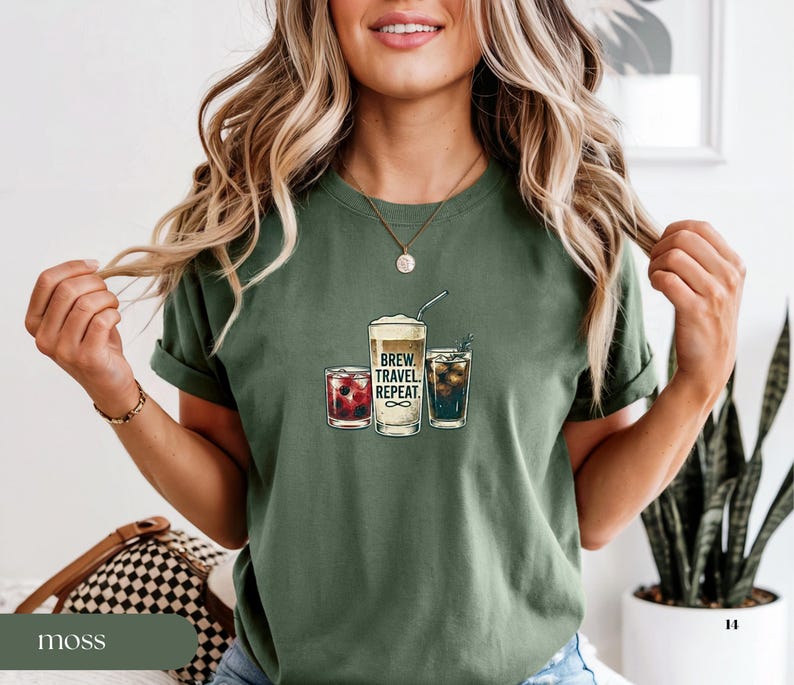 Puede incluir: Camiseta verde musgo con un gr&aacute;fico de tres bebidas: un caf&eacute; espumoso, un c&oacute;ctel rojo y una bebida oscura con hielo. El texto "BREW. TRAVEL. REPEAT." est&aacute; impreso en el caf&eacute;. La modelo lleva un collar dorado.