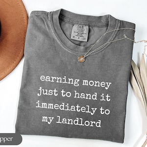May include: A gray Comfort Colors t-shirt with the text "earning money just to hand it immediately to my landlord" in white. A gold necklace and a brown hat are also in the image.