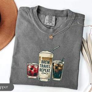 Puede incluir: Una camiseta gris con un gr&aacute;fico de tres bebidas: un c&oacute;ctel, una bebida de caf&eacute; con el texto "BREW. TRAVEL. REPEAT." y una cola. La camiseta tiene cuello redondo y mangas cortas. Un collar dorado est&aacute; drapeado sobre la camiseta.