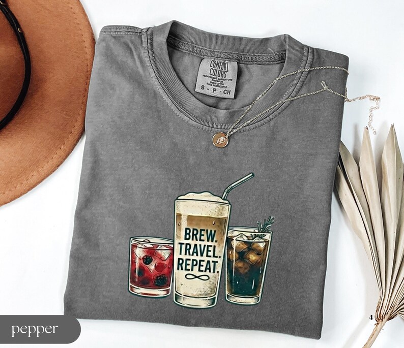 Puede incluir: Una camiseta gris con un gr&aacute;fico de tres bebidas: un c&oacute;ctel, una bebida de caf&eacute; con el texto "BREW. TRAVEL. REPEAT." y una cola. La camiseta tiene cuello redondo y mangas cortas. Un collar dorado est&aacute; drapeado sobre la camiseta.