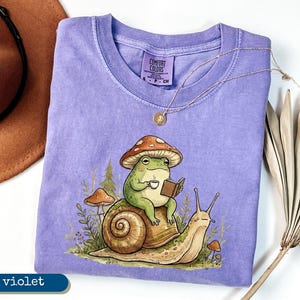 May include: A violet Comfort Colors t-shirt featuring a whimsical illustration of a green frog wearing a mushroom hat, reading a book, and holding a cup of coffee while riding a snail. A gold necklace and a brown hat are also visible.