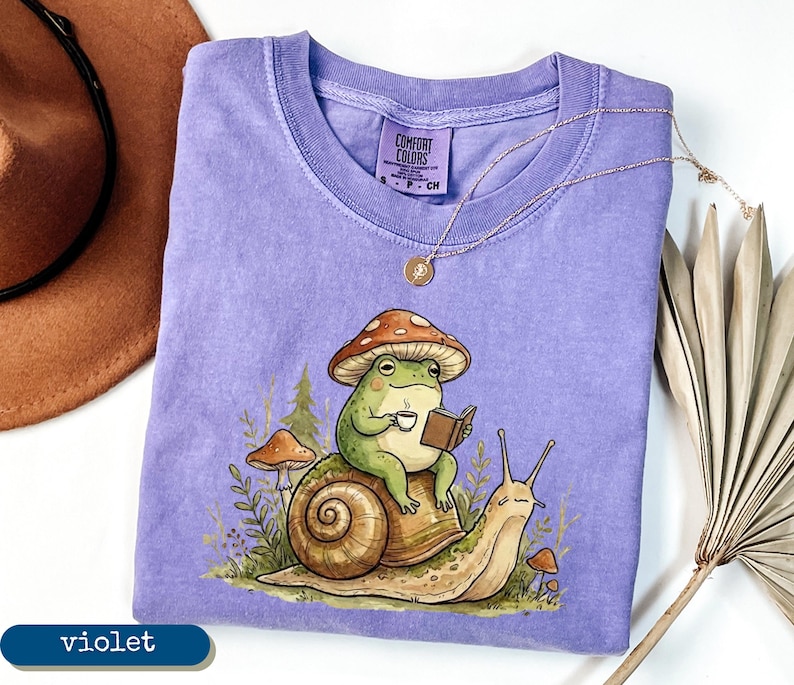 May include: A violet Comfort Colors t-shirt featuring a whimsical illustration of a green frog wearing a mushroom hat, reading a book, and holding a cup of coffee while riding a snail. A gold necklace and a brown hat are also visible.