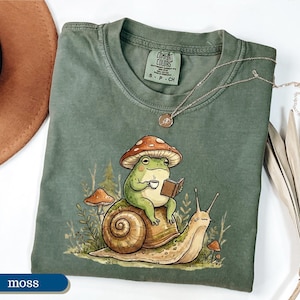 May include: Moss green Comfort Colors t-shirt featuring a whimsical illustration of a frog wearing a mushroom hat, reading a book, and holding a cup of coffee while riding a snail. A gold necklace is draped on the shirt.