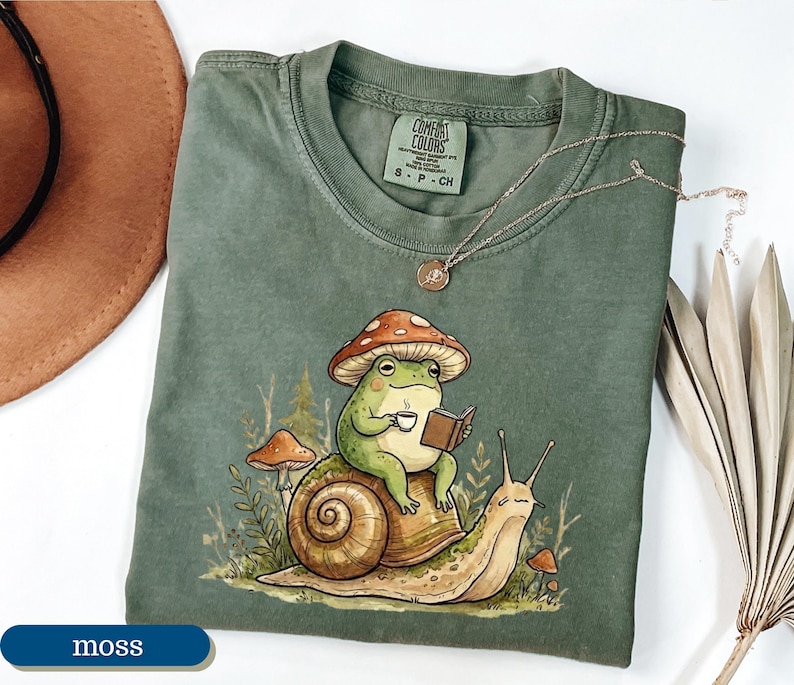May include: Moss green Comfort Colors t-shirt featuring a whimsical illustration of a frog wearing a mushroom hat, reading a book, and holding a cup of coffee while riding a snail. A gold necklace is draped on the shirt.