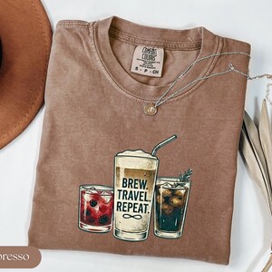 Puede incluir: Una camiseta marr&oacute;n Comfort Colors con un gr&aacute;fico de tres bebidas de caf&eacute; y el texto "Brew. Travel. Repeat." Un collar plateado con un colgante est&aacute; drapeado sobre la camiseta. La camiseta est&aacute; doblada sobre una superficie blanca.