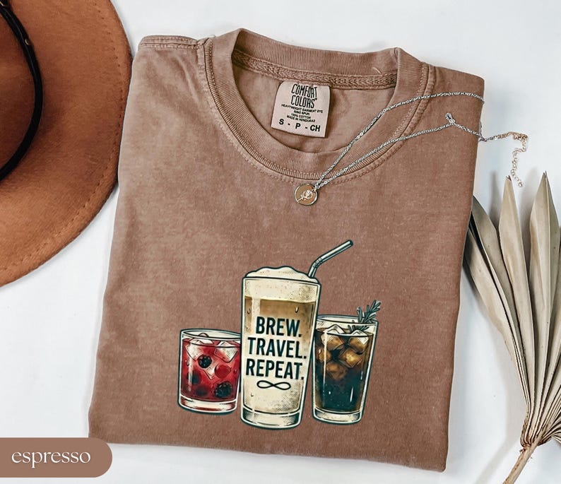 Puede incluir: Una camiseta marr&oacute;n Comfort Colors con un gr&aacute;fico de tres bebidas de caf&eacute; y el texto "Brew. Travel. Repeat." Un collar plateado con un colgante est&aacute; drapeado sobre la camiseta. La camiseta est&aacute; doblada sobre una superficie blanca.