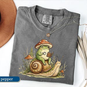 May include: Pepper-colored t-shirt featuring a whimsical illustration of a frog wearing a mushroom hat, reading a book, and holding a cup of coffee while riding a snail. The shirt has a crew neck and short sleeves.