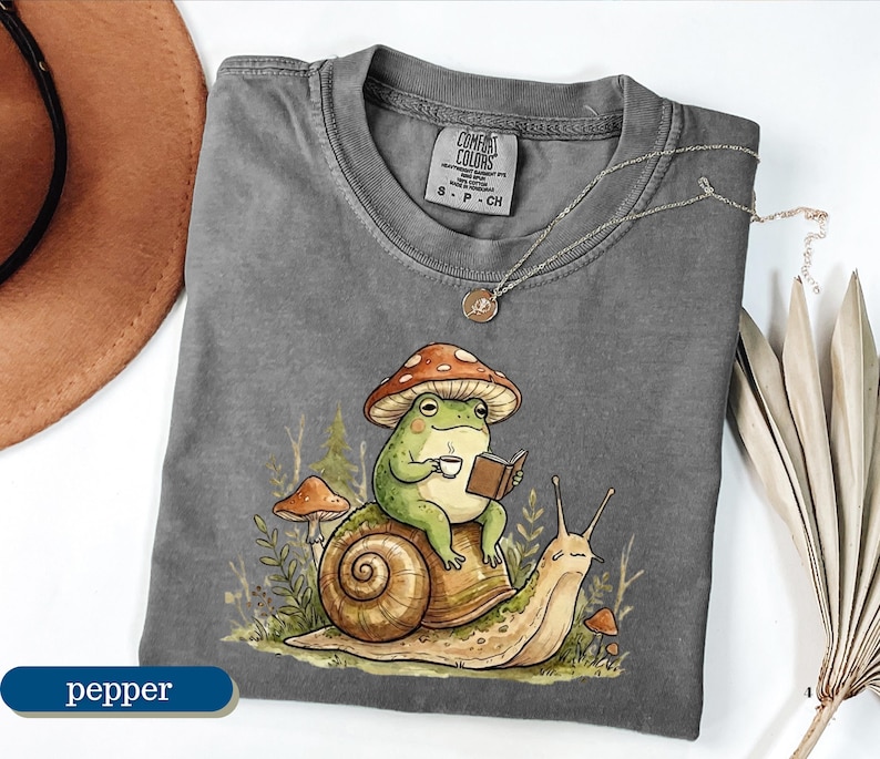 May include: Pepper-colored t-shirt featuring a whimsical illustration of a frog wearing a mushroom hat, reading a book, and holding a cup of coffee while riding a snail. The shirt has a crew neck and short sleeves.