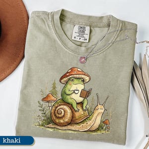 May include: Khaki-colored Comfort Colors t-shirt featuring a whimsical illustration of a frog wearing a mushroom hat, reading a book, and holding a cup of coffee while riding a snail. A silver necklace is draped across the shirt.