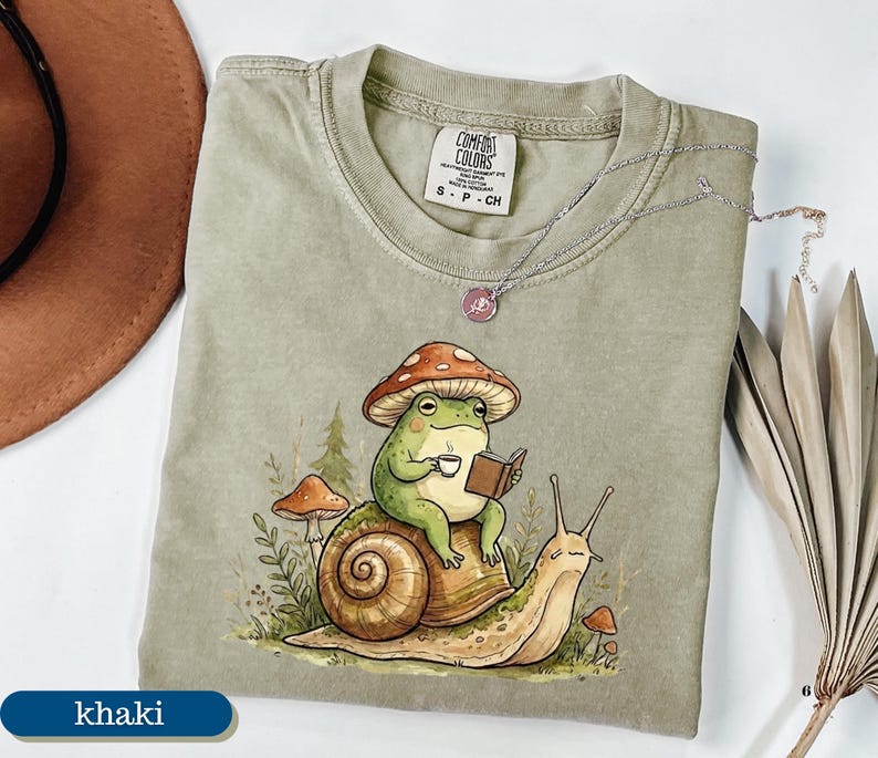 May include: Khaki-colored Comfort Colors t-shirt featuring a whimsical illustration of a frog wearing a mushroom hat, reading a book, and holding a cup of coffee while riding a snail. A silver necklace is draped across the shirt.