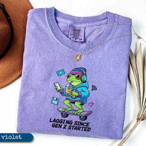 May include: Violet-colored t-shirt with a cartoon frog graphic. The frog is wearing headphones, a hoodie, and holding a drink while riding a skateboard. Text on the shirt reads "LAGGING SINCE GEN Z STARTED". A gold necklace and a brown hat are also visible.