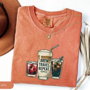 Puede incluir: Una camiseta color &ntilde;ame con un gr&aacute;fico de tres bebidas: un c&oacute;ctel, un caf&eacute; y una cola. El texto "Brew. Travel. Repeat." est&aacute; impreso en el caf&eacute;. Un collar dorado est&aacute; sobre la camiseta.