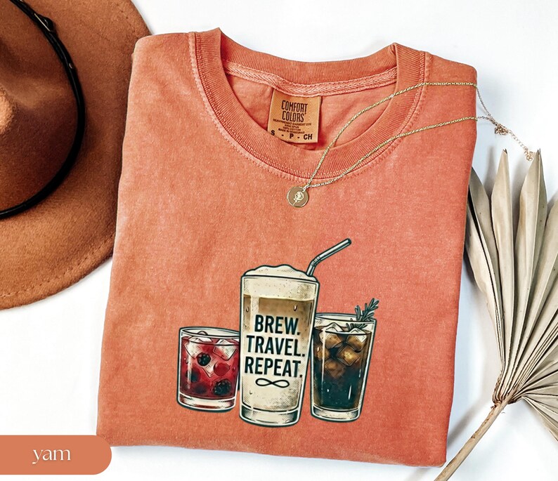 Puede incluir: Una camiseta color &ntilde;ame con un gr&aacute;fico de tres bebidas: un c&oacute;ctel, un caf&eacute; y una cola. El texto "Brew. Travel. Repeat." est&aacute; impreso en el caf&eacute;. Un collar dorado est&aacute; sobre la camiseta.