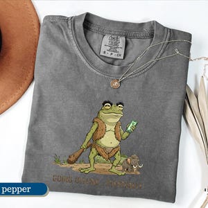 May include: Pepper-colored t-shirt with a humorous cartoon frog design. The frog, dressed in prehistoric attire, holds a smartphone and a club. The text "GOINS ONLINE, BRO" is printed below the frog. A necklace is draped on the shirt.