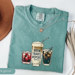 Puede incluir: Camiseta verde claro con un gr&aacute;fico de tres bebidas: un c&oacute;ctel, una bebida de caf&eacute; y un caf&eacute; helado. La bebida de caf&eacute; tiene el texto "Brew. Travel. Repeat." impreso. Un collar plateado con un colgante est&aacute; en la camiseta.