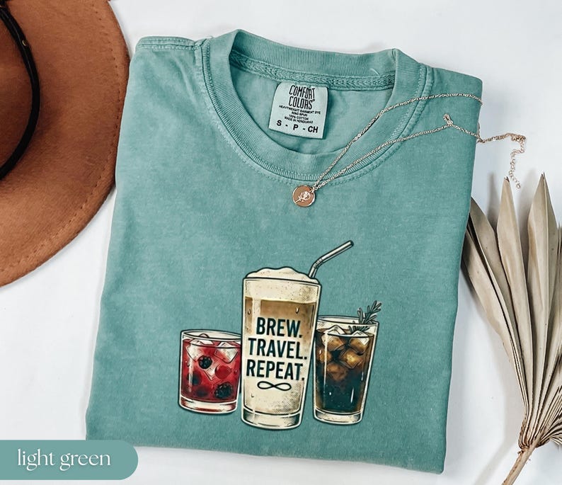 Puede incluir: Camiseta verde claro con un gr&aacute;fico de tres bebidas: un c&oacute;ctel, una bebida de caf&eacute; y un caf&eacute; helado. La bebida de caf&eacute; tiene el texto "Brew. Travel. Repeat." impreso. Un collar plateado con un colgante est&aacute; en la camiseta.