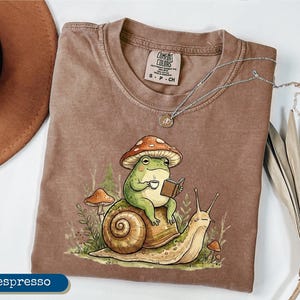 May include: Espresso-colored t-shirt featuring a whimsical illustration of a green frog wearing a mushroom hat, reading a book, and holding a cup of coffee while riding a snail. The shirt has a Comfort Colors tag and a silver necklace.
