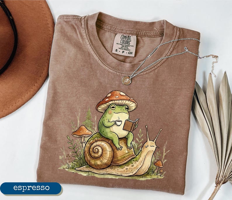 May include: Espresso-colored t-shirt featuring a whimsical illustration of a green frog wearing a mushroom hat, reading a book, and holding a cup of coffee while riding a snail. The shirt has a Comfort Colors tag and a silver necklace.